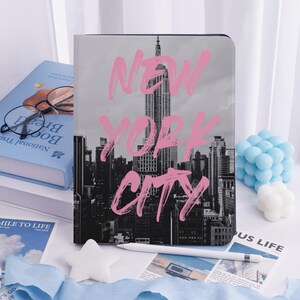 May include: A notebook with a black and white image of the New York City skyline and the words "NEW YORK CITY" in pink. A white stylus and other items are on the white surface.