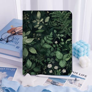 Può includere: Una custodia per tablet con un design botanico dettagliato di varie foglie verdi e fiori bianchi. La custodia è posta su uno sfondo bianco, con uno stilo e un libro visibili. Il design include una varietà di forme e trame di foglie.