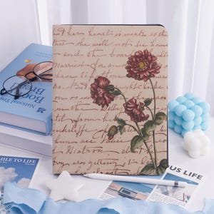 May include: A notebook with a floral design. The cover displays red flowers and handwritten script on a beige background. A white stylus and other items are in the foreground. Other books and accessories are visible.