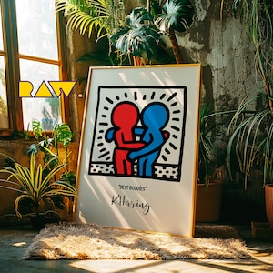 Könnte beinhalten: Ein gerahmter Kunstdruck mit einem Keith Haring-Design von zwei sich umarmenden Figuren, eine rot und eine blau, mit den Worten "Best Buddies" und "KHaring". Das Kunstwerk befindet sich in einem goldenen Rahmen, umgeben von Pflanzen.