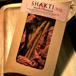 Pode incluir: Uma caixa de cartão castanha com uma janela, contendo varetas de incenso. O rótulo diz "SHAKTI Myrrh Patchouli" com os ingredientes listados. As varetas de incenso são castanhas e repousam sobre papel de seda vermelho. A caixa está numa bandeja de madeira.