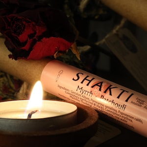 Peut inclure: Une bougie allumée dans un support en bois à côté d'un tube de papier roulé avec le texte "SHAKTI Myrrh Patchouli". Le tube est rose clair avec du texte noir. Une rose rouge foncé est en arrière-plan.