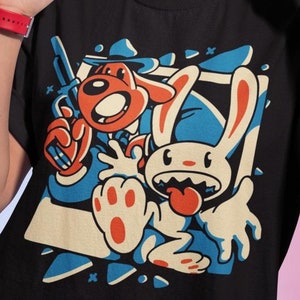 Può includere: Maglietta nera con una grafica a cartoni animati di un coniglio e un cane che indossa un cappello. Il coniglio tiene una pistola e il cane un coltello. La grafica è in uno schema di colori blu, rosso e bianco.