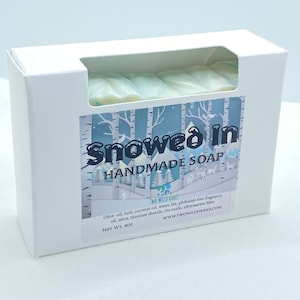 May include: A white box with a window showing a bar of handmade soap. The soap is light blue and white and has a wintery design. The box has the text "Snowed in Handmade Soap" and "Two Wild Hares" on it. The ingredients and website are listed on the box.