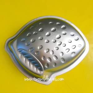 May include: A silver-colored metal cake pan shaped like a strawberry. The pan has a textured surface with small, round indentations to resemble the seeds of a strawberry.