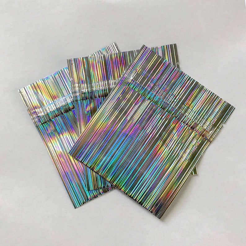 May include: A stack of iridescent silver pouches with a vertical striped pattern. The pouches are made of a thin, flexible material and have a sealed opening.