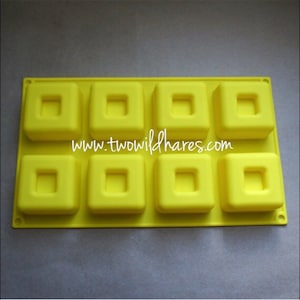 Puede incluir: Molde de silicona amarillo con ocho cavidades cuadradas, cada una con un recorte cuadrado más pequeño en el centro. El molde está diseñado para hacer jabones cuadrados pequeños u otras artesanías. El texto "www.twowildhares.com" está impreso en el molde.