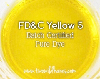 May include: A close-up of a yellow liquid in a clear container. The text "FD&C Yellow 5 Batch Certified Pure Dye" is printed on the container. The website "www.twowildhares.com" is also printed on the container.