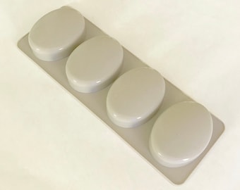 Puede incluir: Un molde de silicona con cuatro cavidades ovaladas para hacer barras de jabón. El molde es gris claro y tiene una superficie lisa.