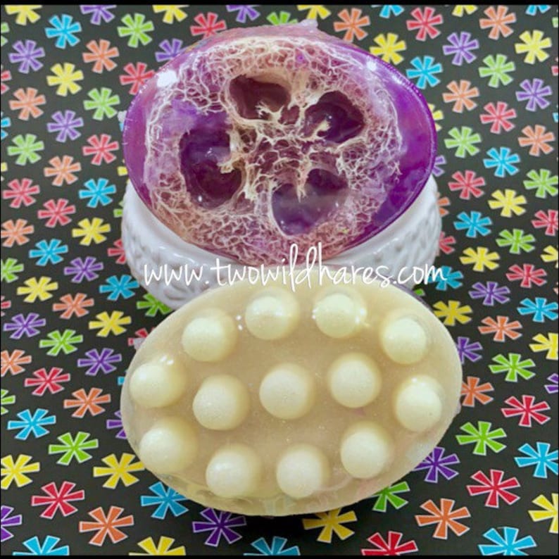 May include: Two oval-shaped bath soaps. The top soap is purple and has a loofah texture. The bottom soap is beige with a textured surface of small white circles. The text "www.twowildhares.com" is visible on the image.