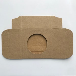 Puede incluir: Una caja de cart&oacute;n marr&oacute;n con un recorte circular en el centro. La caja est&aacute; dise&ntilde;ada para contener un objeto redondo, como un CD o un DVD.
