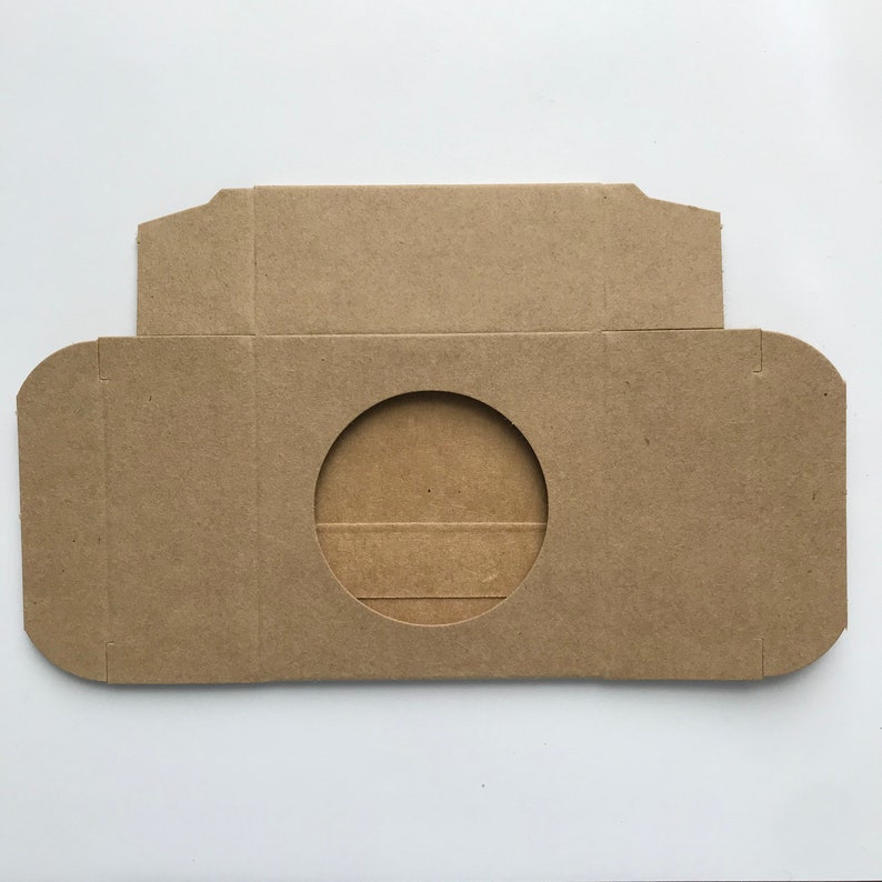 Puede incluir: Una caja de cart&oacute;n marr&oacute;n con un recorte circular en el centro. La caja est&aacute; dise&ntilde;ada para contener un objeto redondo, como un CD o un DVD.