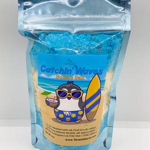 May include: A blue and white pouch with a cartoon penguin wearing sunglasses and holding a surfboard. The pouch contains blue and white bath salts and the text "Catchin' Waves Bubble Salt" is printed on the front. The text "Himalayan pink salt, Dead Sea salt, sodium bicarbonate, lathanol, dendritic salt, tapioca starch, phthalate free fragrance oil, FD&C blue 1, FD&C yellow 5" is printed on the back. The text "Net Wt 7oz" is printed on the bottom. The text "www.Twowildhares.com" is printed on the bottom.