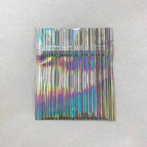 May include: A silver iridescent plastic bag with a heat-sealed opening. The bag is made of thin, flexible plastic and has a holographic finish.