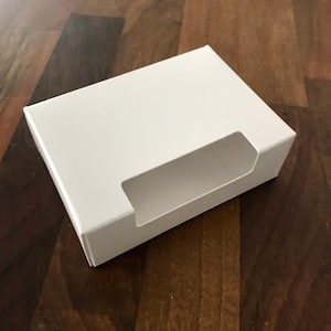 May include: A white cardboard box with a rectangular opening on the front. The box is designed to hold small items.