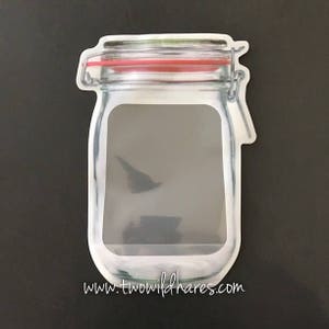 May include: A clear plastic reusable food storage bag shaped like a glass jar with a red lid and a zipper closure. The bag has a white background with the text "www.twowildhares.com" printed on the bottom.