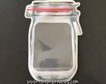 May include: A clear plastic reusable food storage bag shaped like a glass jar with a red lid and a zipper closure. The bag has a white background with the text "www.twowildhares.com" printed on the bottom.