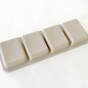 Puede incluir: Un molde de silicona gris con cuatro cavidades cuadradas. El molde está diseñado para hacer objetos pequeños de forma cuadrada.