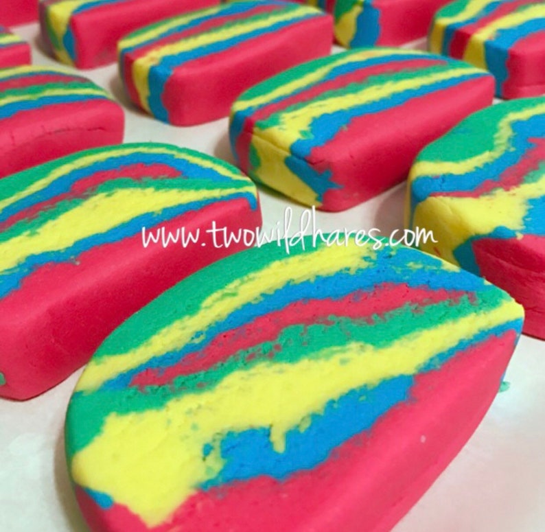 Puede incluir: Primer plano de barras de jab&oacute;n coloridas y en espiral. El jab&oacute;n es rojo, amarillo, azul y verde. El jab&oacute;n tiene forma de rect&aacute;ngulo y una superficie lisa y brillante. El jab&oacute;n est&aacute; dispuesto en una fila sobre una superficie blanca. El texto "www.twowildhares.com" es visible en la imagen.