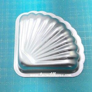 May include: A silver metal baking pan shaped like a seashell with a scalloped edge. The pan has a ribbed texture and is designed for baking cakes or other desserts.