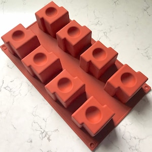 May include: A brown silicone mold with seven square cavities, each with a round indentation in the center. The mold is designed for making small cakes or other treats.