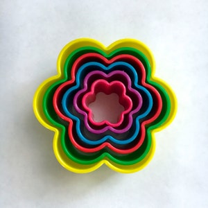 Puede incluir: Conjunto de cinco cortadores de galletas con forma de flor en un arcoíris de colores, incluyendo amarillo, verde, rojo, azul y morado.