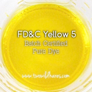 May include: A close-up of a yellow liquid in a clear glass jar. The text "FD&C Yellow 5 Batch Certified Pure Dye" is printed on the jar. The website "www.twowildhares.com" is printed at the bottom of the jar.