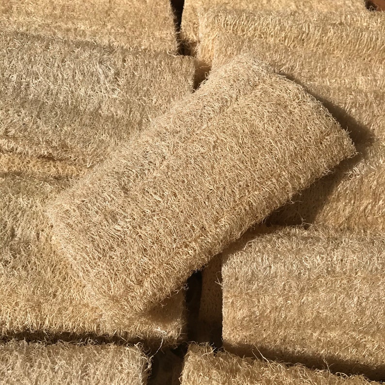 May include: A collection of beige loofah sponges, rectangular in shape, with a natural, fibrous texture. One sponge is positioned on top of the others, highlighting its form. These eco-friendly bath accessories are made from dried gourds.