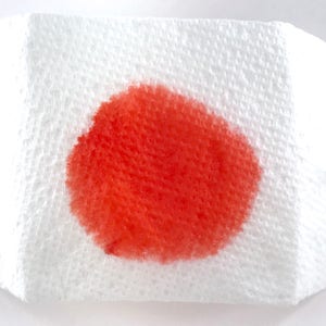 May include: A close-up of a white paper towel with a bright red stain in the center.