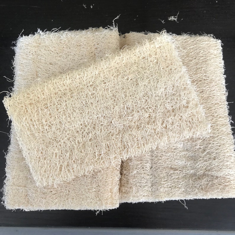 May include: A stack of rectangular loofah sponges in a natural, off-white colour. The sponges have a fibrous, open weave texture. These eco-friendly sponges are suitable for use in the bath or kitchen. The sponges are stacked on a dark surface.