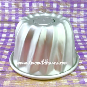 Op de afbeelding: Zilverkleurige metalen bundt cakevorm met een geribbeld ontwerp. De vorm heeft een losse bodem en een website-adres dat aan de zijkant is gedrukt: www.twowildhares.com.