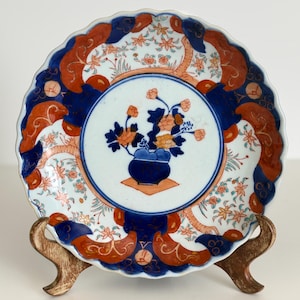 May include: A decorative ceramic plate with a scalloped edge, featuring a central floral design in a blue vase. The plate has a blue and orange border with floral and scroll patterns. The plate is displayed on a gold-toned stand.