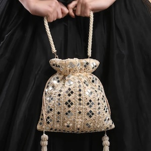 May include: A beige, embellished drawstring bag with a pearl-beaded handle and decorative tassels. The bag is covered in small, square mirrored accents and is held against a black background.