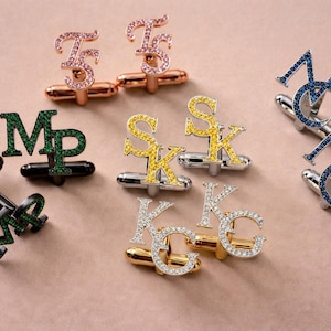Puede incluir: Un surtido de gemelos personalizados, cada uno con iniciales en varios colores y adornos de gemas. Los gemelos son de oro rosa, plata y negro, con iniciales en gemas rosas, amarillas, azules, verdes y transparentes. El fondo es rosa suave.