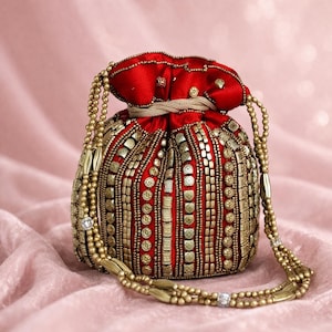 May include: A red pouch bag, embellished with gold-coloured beads and a drawstring closure. The bag features a beaded strap and is set against a pink, silky backdrop. This bag is likely designed for carrying small items.