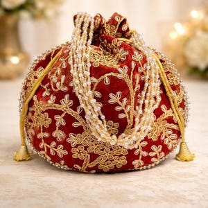 May include: A round, red velvet pouch with gold floral embroidery and a beaded pearl strand. The drawstring closure has gold tassels. The pouch is embellished with small silver beads around the edges.