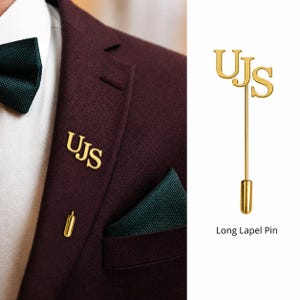 Puede incluir: Un prendedor largo dorado con las letras "UJS" en una fuente serif. El prendedor se muestra en una blazer burdeos, con una pajarita y un pañuelo de bolsillo verde oscuro. El prendedor mide aproximadamente 7.6 cm de largo.