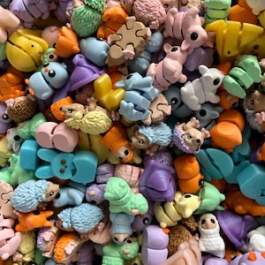 May include: A colorful assortment of miniature animal figurines in various shapes and colors. The collection includes hedgehogs, bunnies, and other creatures in shades of blue, green, orange, and purple. Some are puzzle pieces. The figurines are made of a smooth material.