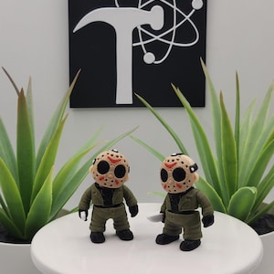 May include: Two small figurines wearing tan hockey masks with red markings and black eye holes. Each figure wears a green jacket, green pants, and black boots. One figure holds a small white knife in its right hand, while the other holds it in its left.