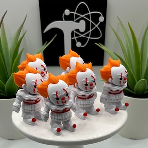 May include: A collection of small, white clown figurines with orange hair and red accents. The figurines are arranged on a white, round surface, with green plants in white pots on either side. A black sign with a white logo is in the background.