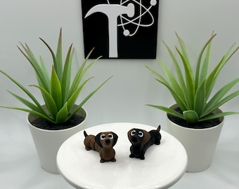 May include: Two dachshund figurines, one brown and one black, sit on a white pedestal. The figurines are detailed with small eyes and ears. Two potted green plants flank the pedestal. A black and white sign is in the background.