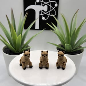 May include: Three brown alpaca figurines with black eyes and noses. The figurines are sitting on a white cake stand.