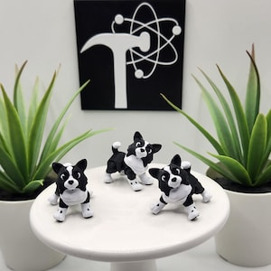 May include: Three small black and white dog figurines with pointed ears and white chest markings are positioned on a white circular pedestal. Behind the pedestal, a black square sign displays a white hammer icon overlaid on a white atom symbol.