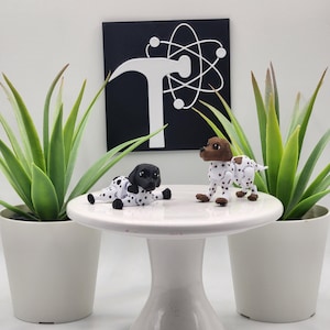 May include: Two Dalmatian dog figurines, one lying down and one standing, are displayed on a white pedestal. The dogs have black and white spots. The pedestal is in front of a black sign with a white graphic and two potted plants.