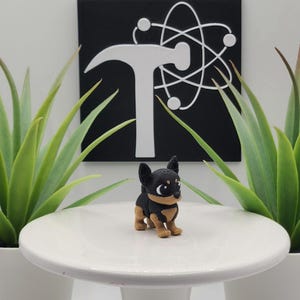 May include: A small, black and tan Chihuahua figurine stands on a white pedestal. The figurine is in front of a black square with a white hammer and atom symbol. Two potted green plants flank the pedestal.