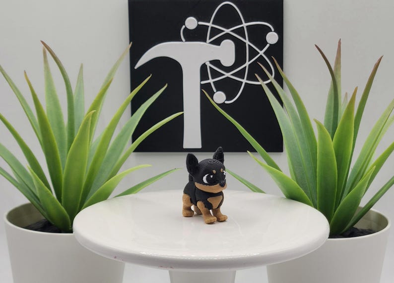 May include: A small, black and tan Chihuahua figurine stands on a white pedestal. The figurine is in front of a black square with a white hammer and atom symbol. Two potted green plants flank the pedestal.