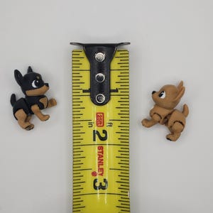 May include: Two small, detailed Chihuahua figurines, one black and tan, the other tan, are positioned on a white surface. A yellow Stanley measuring tape is between them, showing measurements in inches. The figurines are approximately 1 inch in height.
