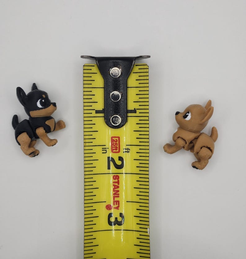 May include: Two small, detailed Chihuahua figurines, one black and tan, the other tan, are positioned on a white surface. A yellow Stanley measuring tape is between them, showing measurements in inches. The figurines are approximately 1 inch in height.