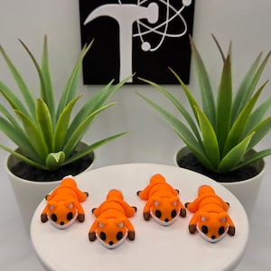 May include: Four orange fox figurines with white bellies and brown accents are displayed on a white cake stand. Two potted green plants flank the figurines. A black sign with a white logo is in the background.