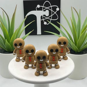 May include: Five brown gingerbread man figurines with black eyes and red button details are arranged on a white cake stand. The figurines are in front of two potted green plants and a black sign with a white graphic.